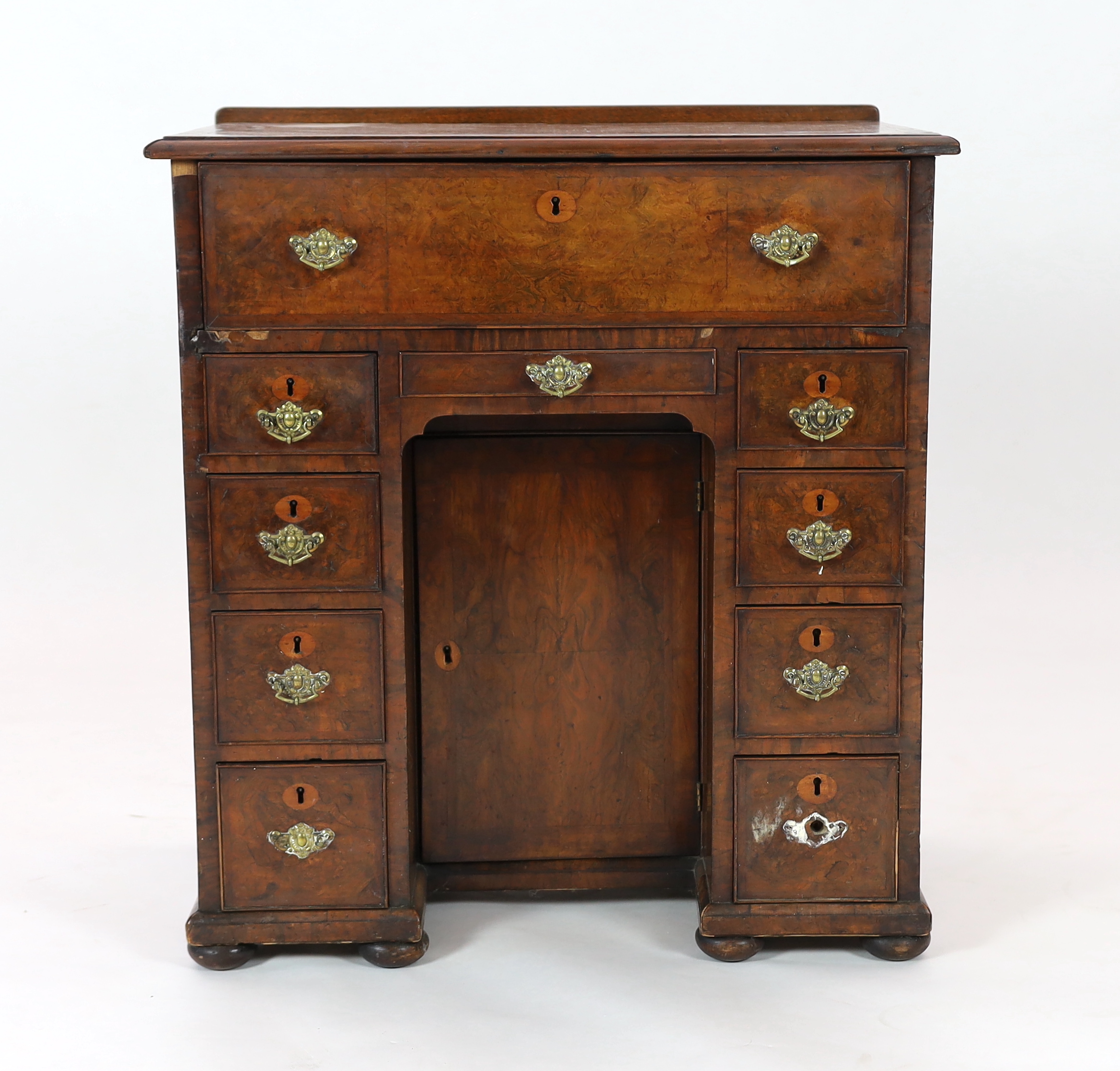 An early 18th century walnut kneehole secretaire desk, with quarter veneered top and fall front drawer opening to reveal pigeon holes and drawers, over eight further drawers surrounding a central recessed cupboard, on pa